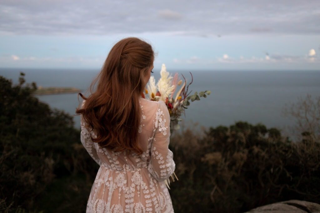 Bride in a lace dress holding a dried-flower bouquet, looking out to sea