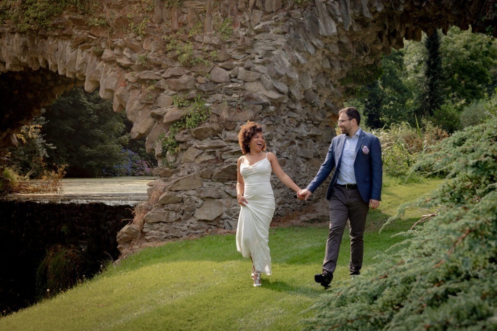 Bride and groom walking hand in hand past a stone arch